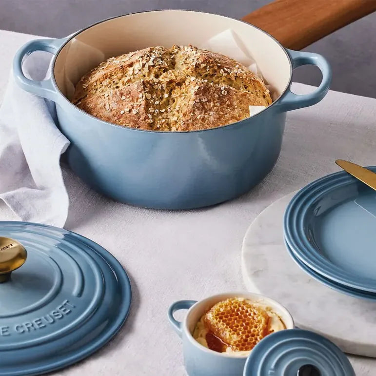 A loaf of bread baked in the casserole, on a table with plates and the lid resting beside it, in chambray blue