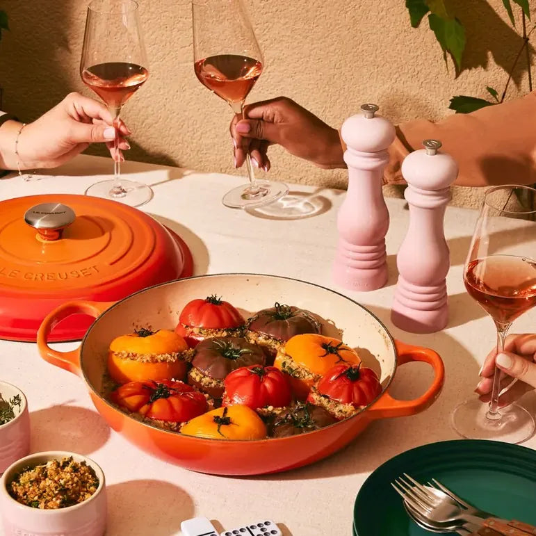 A shallow casserole of stuffed tomatoes on a laid table, the lid resting beside it and a hand lifting a glass